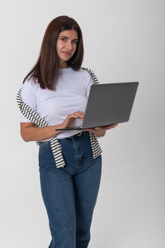 Young adult woman in casual wear standing in studio, confidently typing on a laptop while looking at camera, conveying modern remote work, digital communication and networking