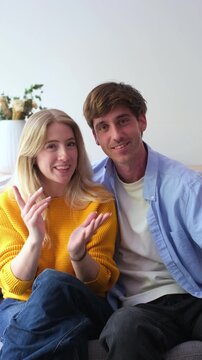 Millennial boyfriend and girlfriend sitting together on a couch, smiling and talking to the camera while recording a vlog or making a video call to family in their modern apartment