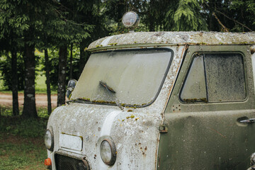 Fototapeta premium Vintage green and white delivery vehicle covered in moss and dirt, parked in a forested area with tall trees and a dirt path visible in the background