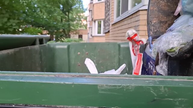 Close-up of green plastic dumpster filled with household waste bags against blurry background of residential buildings. Dumping waste into public dumpster. Infrastructure and work of public utilities