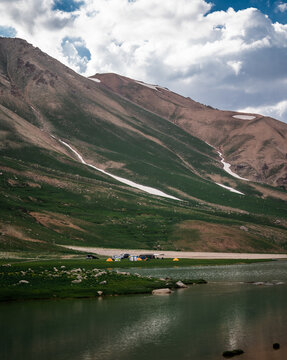 View of Arashan Lake with colorful tents and vehicles on the grassy shore, set against dramatic mountain slopes with patches of snow in Pop District, Namangan Region, Uzbekistan.