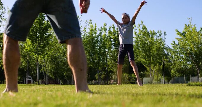 Father and son play soccer barefoot on green grass at sunset. Child celebrates successful kick with raised arms and joyful jump while dad smiles during family game in park.