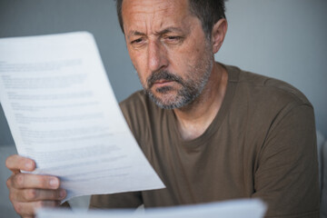 Fototapeta premium Middle-aged man studies a printed document intently, hand on chin and furrowed brow conveying worry, concentration and contemplation while reading important paperwork at home.