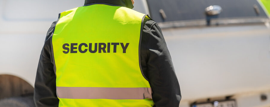 Rear view of security officer wearing yellow reflective vest labeled SECURITY while observing people in sunny urban setting