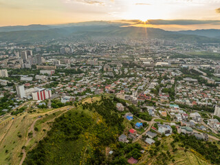 Obraz premium A stunning high-angle aerial drone shot overlooking the expansive cityscape of Tbilisi, Georgia, during a vibrant sunset. The composition highlights a rolling green and golden hill with a dirt path