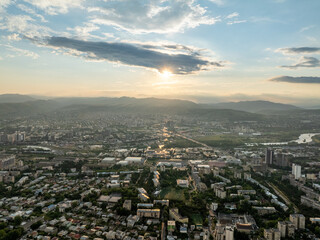 Fototapeta premium A stunning high-angle aerial drone shot overlooking the expansive cityscape of Tbilisi, Georgia, during a vibrant sunset. The composition highlights a rolling green and golden hill with a dirt path