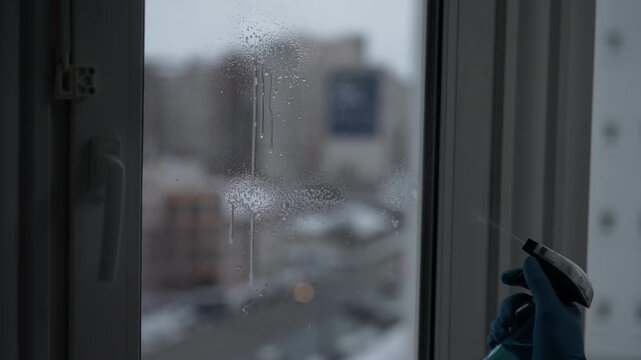 Professional cleaning service worker spraying cleaning solution onto window and then wiping it with microfiber cloth, revealing blurry city view with traffic outside on cloudy day, slow motion.