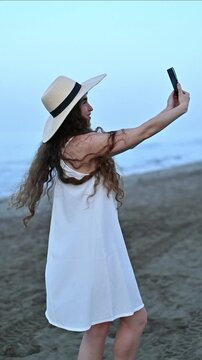 Young Happy Woman Recording Vertical Selfie Video While Walking on Sunny Beach, Smiling Travel Content Creator Filming Authentic UGC Social Media Lifestyle, Summer Vacation by the Ocean Coast
