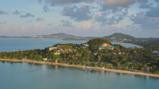 Tropical island coastline with green hills, luxury villas resorts, palm trees, extending along a sandy beach and blue ocean water under cloudy sky. Travel Samui, Thailand. Aerial view drone flight