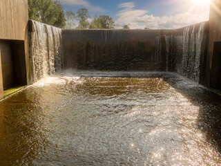 Water cascades down concrete dam into calm pond below. Sunlight reflects on rippling water surface. Trees stand behind dam in background. Scene shows man-made water feature outdoors © mark_gusev