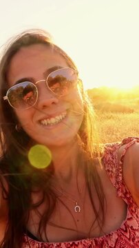 Beautiful young woman with sunglasses smiling and laughing while taking a selfie video in a golden field at sunset, her hair blowing in the wind during a carefree summer evening