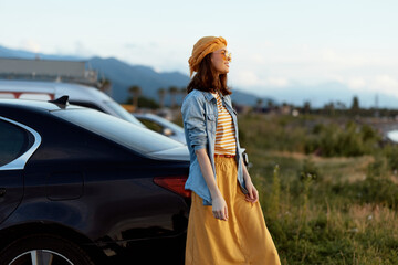Obraz premium A woman stands beside a sleek car in a rural landscape, wearing a striped top, denim shirt, and mustard dress with a sun hat. Casual lifestyle moment focused on outdoor fashion and calm atmosphere.