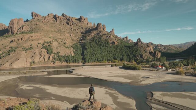 Wide shot of a hiker overlooking the confluence of the Limay and Traful rivers, set against rocky mountain terrain and a forest in Patagonia, Argentina.