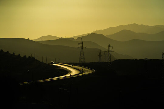 View of a winding highway at sunset with electricity pylons and layered mountain silhouettes in the background in Pop District, Namangan Region, Uzbekistan.