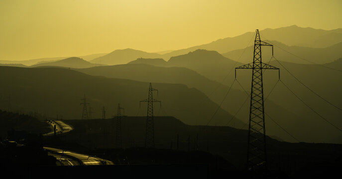 View of power lines silhouetted against layered mountain ranges during golden hour with a winding road in the foreground in Pop District, Namangan Region, Uzbekistan.