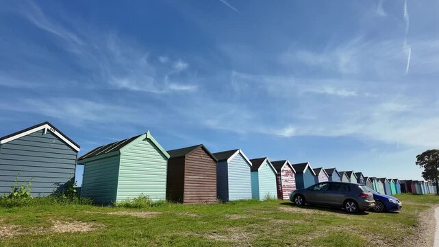 Hampshire England UK. 12.06.2026. Video. A row of vibrant, pastel-colored beach huts stands on a grassy shore beneath a clear blue sky dotted with thin, wispy clouds