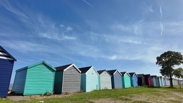 Hampshire England UK. 12.06.2026. Video. A row of vibrant, pastel-colored beach huts stands on a grassy shore beneath a clear blue sky dotted with thin, wispy clouds