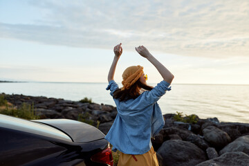 Obraz premium Woman in denim shirt and straw hat stands by rocky coast at sunset, joyful mood, arms raised, travel lifestyle scene near seaside rocks and car