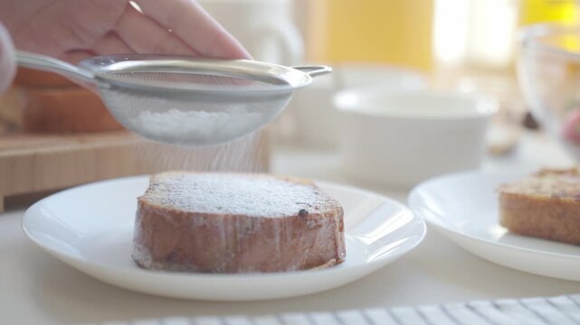 Dusting French toast with powdered sugar, using small sieve over cooked slice on plate. Concept of breakfast and homemade dessert