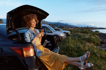 Obraz premium Cheerful woman outdoors, smiling while sitting in a car trunk, warm sunset glow, casual denim jacket over a yellow dress, relaxed lifestyle moment