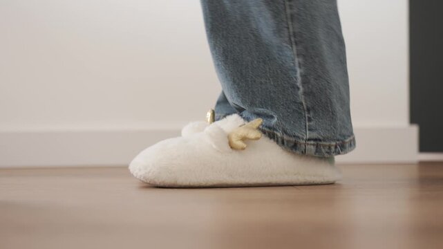 Woman in striped socks puts on white fluffy slippers and walks across wooden floor. Concept of home comfort and casual indoor footwear