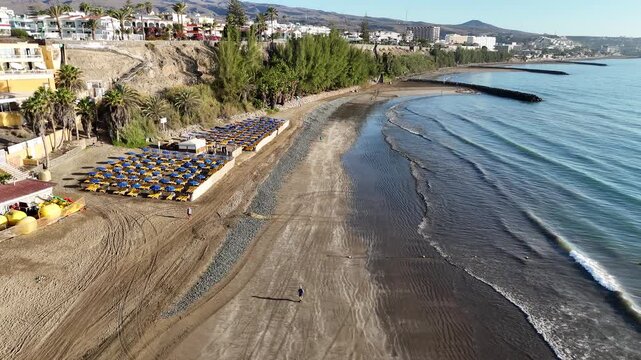 Aerial panorama flight over beach with tourists at Atlantic ocean towards cliff coast of Playa del Ingles with hotels and hills on Canary Island Gran Canaria, Spain