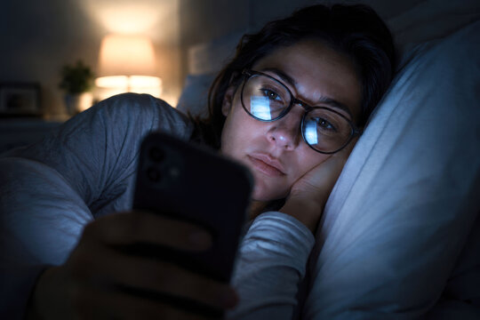 Young woman lying awake in bed using smartphone late at night with screen reflection in eyeglasses showing digital addiction insomnia anxiety sleep problems technology dependence and mental health
