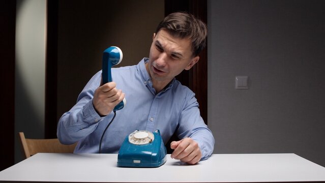 Man in a blue shirt sitting at a table, holding a vintage blue rotary phone receiver, expressing confusion and misunderstanding about outdated communication technology