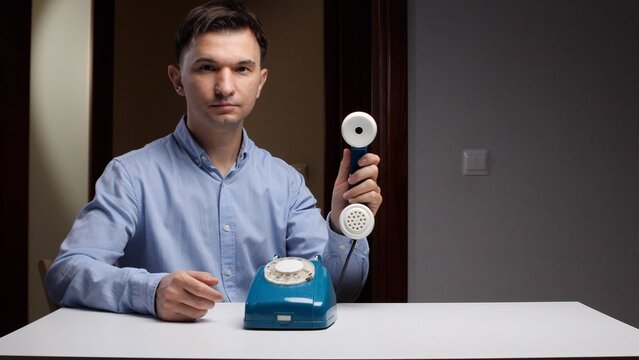 Young man in a blue shirt holding a retro telephone receiver, looking directly at the camera while sitting at a white table, communicating with an outdated device
