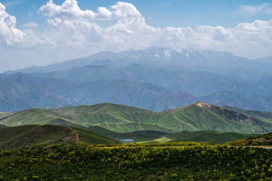View of Arashan ko'li nestled among rolling green hills and distant snow-capped mountains under a cloudy blue sky in Pop District, Namangan Region, Uzbekistan.