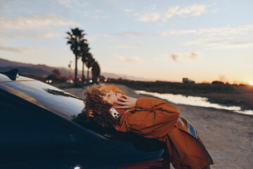 Obraz premium Woman in lifestyle portrait, curly hair leaning on car hood at sunset near ocean shoreline, wearing orange jacket, hands touching hair, warm golden light, scenic road background, outdoors