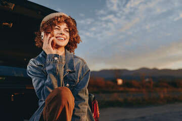 Obraz premium Woman lifestyle moment, smiling woman wearing denim jacket, holding phone to ear, curly hair, warm sunset sky, outdoors in evening air, relaxed casual travel mood, city background blur