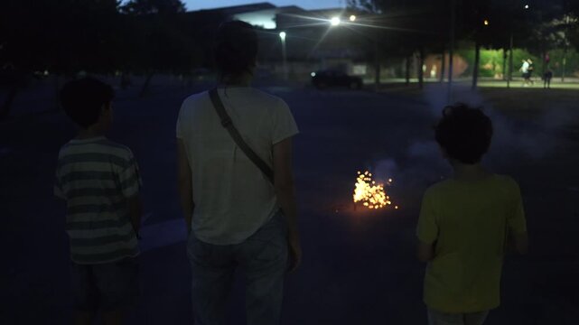 Mother and two sons watching a firework exploding on the asphalt ground at night. Family enjoying the sparks and bright lights of a ground firecracker in a city park