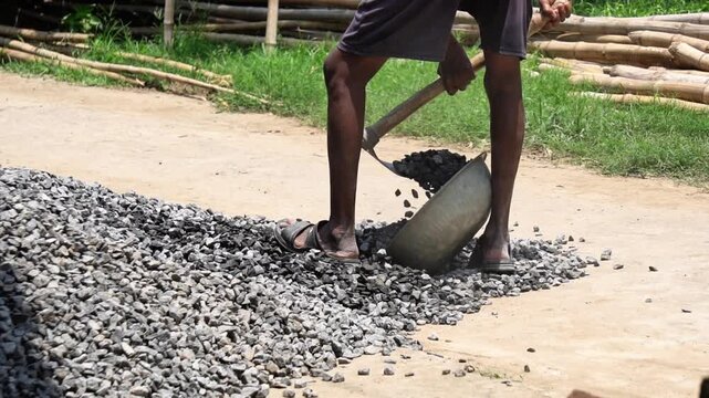 Indian Laborer Shoveling Crushed Stones Into Metal Basket on Road Construction Site. Cinematic Manual Labor Scene of Indian Man Filling Iron Pan with Stones.