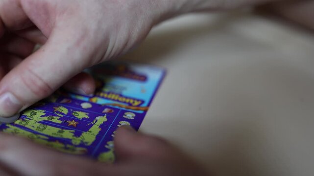 Close up of a hand scratching a colorful lottery ticket with a coin searching for a winning combination dreaming of a jackpot and testing personal luck
