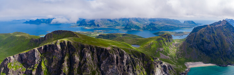 Fototapeta premium Aerial view of Ryten mountain cliffs above turquoise fjords and sandy coves in Lofoten Islands, Norway. Low clouds and soft light suggest a cool late summer evening.
