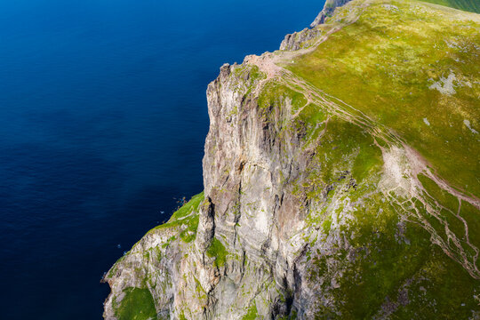 Aerial view shows Ryten cliff in Lofoten, Norway, with grassy plateau, braided hiking paths, and drop to the Norwegian Sea in daylight, strong top down composition.