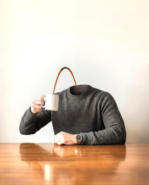 Whimsical personification of a morning beverage, this coffee head figure sits at a wooden table. The ceramic cup, or muggy, replaces the head of a person in a textured grey sweater.