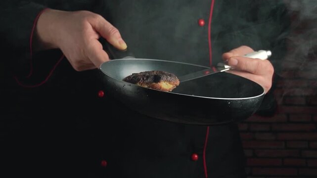 Chef in black coat flips cooked meat in frying pan, showcasing culinary skills in a smoky kitchen with brick wall background
