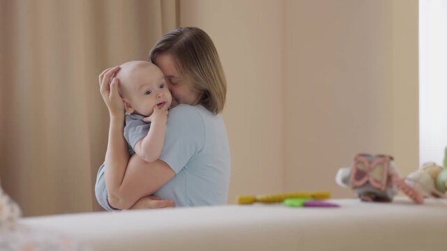 Caucasian mother cuddles baby after playtime on bed, toys scattered on mattress, cozy blanket and pillow, postplay calm, gentle snuggle, eye contact and quiet bond