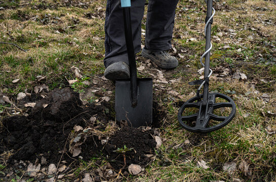 A person stands in a park using a metal detector. They have dug a hole in the ground with a shovel and search for items. The grass and leaves are scattered around