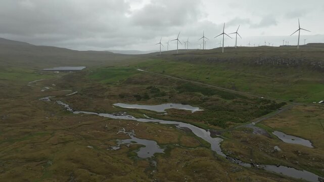 Aerial drone footage panning left over a rural landscape with wind turbines, a winding stream of river Sand&aacute;, and small lakes on the Faroe Islands under a cloudy sky.