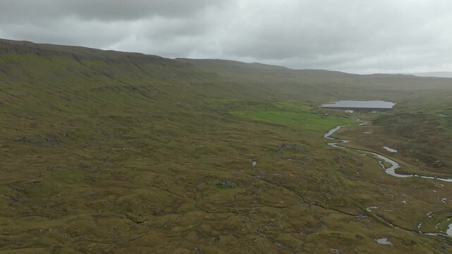 An aerial drone shot pushing in on a remote, lush landscape with hills, a winding stream of river Sand&aacute;, and a tranquil lake on the Faroe Islands under a cloudy sky.