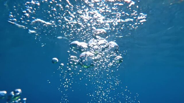 Slow motion water bubbles rise in clear blue water with light reflections from above