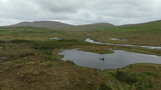 Aerial drone shot panning left over a lush, remote valley on the Faroe Islands, featuring the winding river Sand&aacute;, the tranquil Grunnatj&oslash;rn lake, and a distant village under cloudy skies.