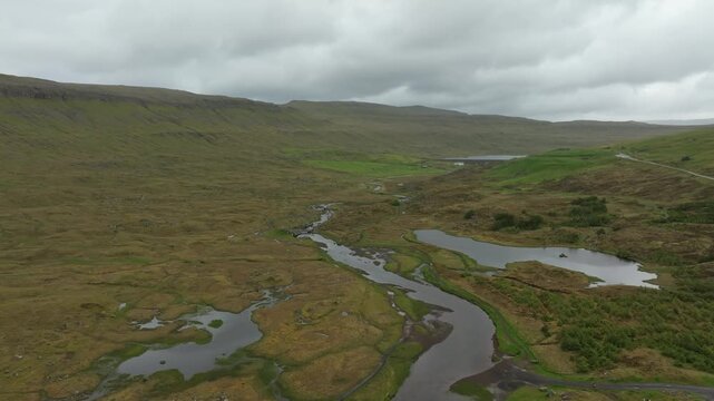 An aerial push in drone shot reveals a vast, rugged green valley on the Faroe Islands. The winding river Sand&aacute;, several small lakes, and remote buildings dot the cloudy landscape.