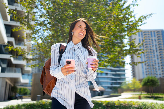 A woman walks outdoors with two coffee cups in her hands. She is smiling and looking at her phone while wearing a backpack. The scene is in a city with tall buildings and greenery.