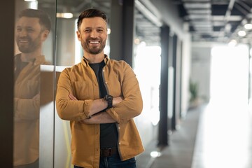 Fototapeta premium Smiling Young Caucasian Man With Beard Standing In Modern Office Space, Arms Crossed, Exuding Confidence And Professionalism. Bright Atmosphere Reflecting A Business Concept.