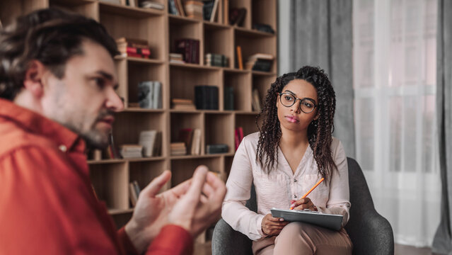 Focused Black Female Therapist Engaging With Male Client During Therapy Session At Office, Both Attentive And Professional. Reflecting On Mental Health And Wellness Concept.
