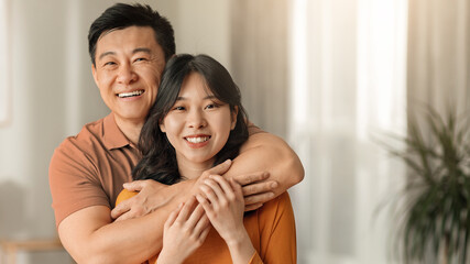 Obraz premium Joyful Asian Couple, Man And Woman, Smiling And Embracing Each Other In Their Bright Living Room At Home, Symbolizing Love And Togetherness, Family Concept.